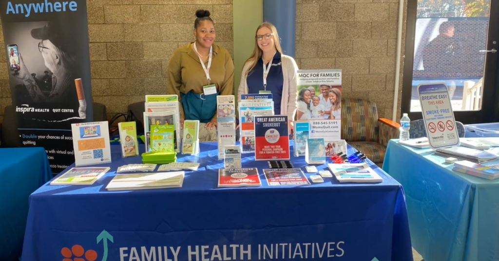 Two staff members smiling from behind a Family Health Initiatives containing information.